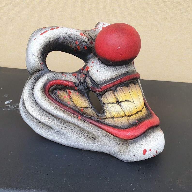 May include: A white ceramic clown mask with a red nose and a wide, toothy grin. The mask has a distressed paint finish and red and yellow accents.