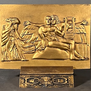 Könnte beinhalten: Eine goldfarbene Wandplakette mit einer Reliefskulptur von drei Figuren. Die zentrale Figur ist ein nackter Mann mit einer Leier, flankiert von zwei drapierten Frauenfiguren. Die Plakette ist auf einem Holzsockel mit einem verzierten Design montiert.