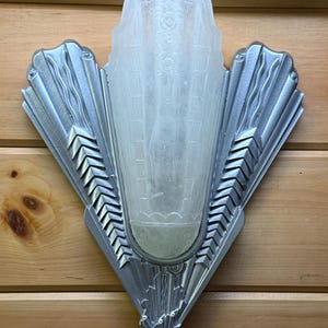 May include: Art Deco style wall sconce with a frosted glass shade and a silver-colored metal frame. The frame features a fan-shaped design with geometric details and a pointed bottom. The glass shade has a vertical pattern.