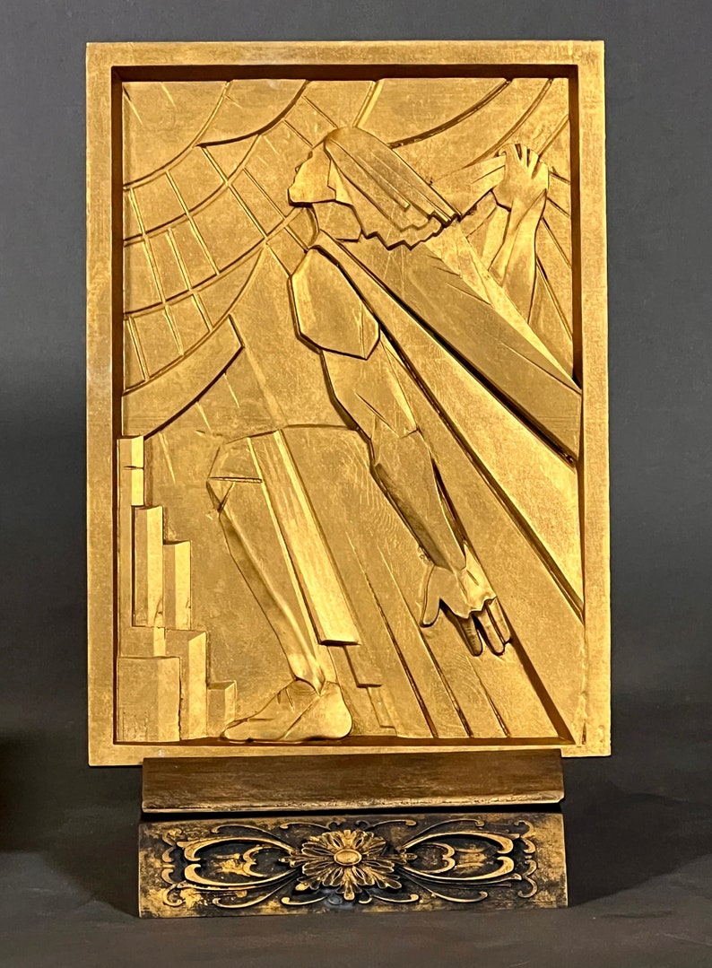K&ouml;nnte beinhalten: Eine goldfarbene Holzskulptur einer Person, die auf einer Plattform steht. Die Skulptur ist im Stil des Art Deco gehalten und weist geometrische Formen auf.