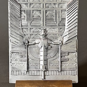 Op de afbeelding: Een zilveren metalen sculptuur van een gevleugelde figuur met een kroon en een staf in elke hand. Het beeld staat op een gouden metalen standaard met een sierlijk ontwerp.