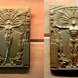 Könnte beinhalten: Zwei Reliefplatten im Art-Deco-Stil, eine mit einer weiblichen Figur und die andere mit einer männlichen Figur, beide mit ausgestreckten Armen. Die Tafeln sind aus goldfarbenem Metall gegossen und vor einem Holzgrund angebracht.