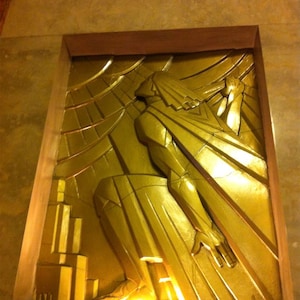 K&ouml;nnte beinhalten: Eine goldfarbene Reliefskulptur einer Figur mit ausgestreckten Armen vor einem Hintergrund aus geometrischen Formen.
