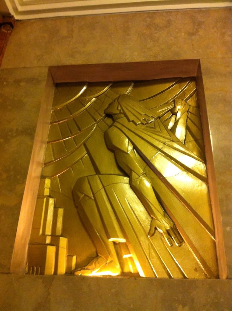 K&ouml;nnte beinhalten: Eine goldfarbene Reliefskulptur einer Figur mit ausgestreckten Armen vor einem Hintergrund aus geometrischen Formen.