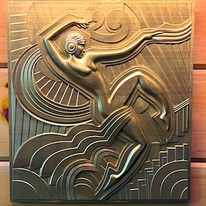 Op de afbeelding: Een goudkleurige metalen wandsculptuur die een gestileerde vrouwelijke figuur in een dynamische pose voorstelt. De figuur is omgeven door geometrische vormen en lijnen, waardoor een gevoel van beweging en energie ontstaat.
