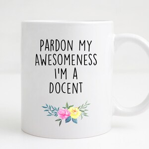 May include: White ceramic coffee mug with black text that reads "Pardon my awesomeness I'm a docent" and a floral design with pink and yellow flowers.