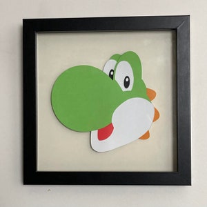 May include: A framed paper craft of Yoshi, a green dinosaur-like character from the Super Mario video game series. Yoshi has a large, green nose and a white body with a red tongue sticking out.