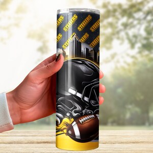 Puede incluir: Un vaso de acero inoxidable negro y dorado con un casco de f&uacute;tbol americano y un gr&aacute;fico de f&uacute;tbol. El vaso tiene un fondo negro con un patr&oacute;n repetido de la palabra "Steelers" en letras doradas.
