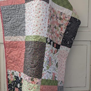 May include: A patchwork quilt featuring a range of floral patterns in shades of pink, green, and black. The quilt is made up of squares and rectangles, and the fabric is quilted with a simple stitch.