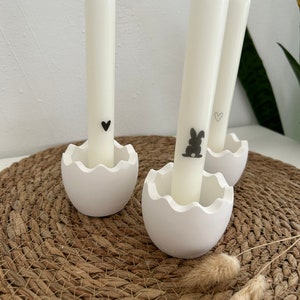 Könnte beinhalten: Drei weiße Kerzen in eiförmigen Haltern mit schwarzen Hasen- und Herzmotiven. Die Kerzenhalter stehen auf einer geflochtenen braunen Matte.