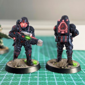 May include: Two miniature soldier figurines in black armor with red and green highlights. One is viewed from the front, holding a futuristic rifle. The other is viewed from the back, with a glowing emblem on the backpack. Both stand on a circular base.