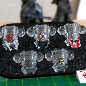 Puede incluir: Cinco piezas de armadura en miniatura de plástico transparente con varios diseños de emblemas. Los emblemas incluyen cruces rojas, calaveras y otros símbolos, colocados sobre una superficie negra texturizada. Las piezas de armadura son probablemente para juegos de mesa o modelismo.