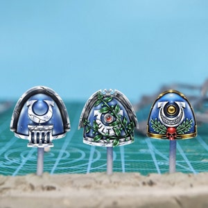 May include: Three blue and silver miniature helmet designs. Each helmet features unique emblems and details, including a laurel wreath, a skull, and architectural elements. The helmets are mounted on gray rods against a light blue background.