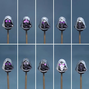 Könnte beinhalten: Eine Sammlung von zehn Miniatur-Gasmaskenhelmen mit Kapuzendesign und violetten Details. Die Helme sind auf Holzstäben vor blauem Hintergrund montiert. Die Helme haben eine metallische Oberfläche und ein futuristisches Aussehen.