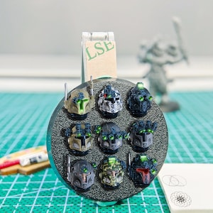May include: A collection of nine miniature, painted sci-fi helmet models, each with unique color schemes and details. The helmets are arranged on a circular base, showcasing various designs with green and red accents. The background includes a miniature figure and a cutting mat.
