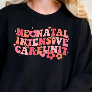 May include: A black sweatshirt with a pink and red graphic design that says "Neonatal Intensive Care Unit" with a heart and flowers.