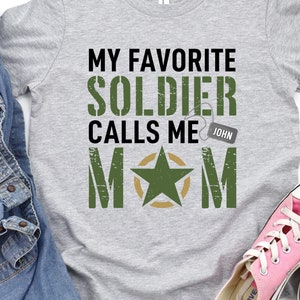 May include: A grey t-shirt with the text "MY FAVORITE SOLDIER CALLS ME MOM" in green and black. A denim jacket and pink trainers are next to the shirt. The shirt features a military star graphic.
