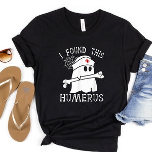 May include: Black t-shirt with the text "I FOUND THIS HUMERUS" and a cartoon ghost wearing a nurse's cap and holding a bone. The shirt is paired with denim shorts, sunglasses, and flip-flops.