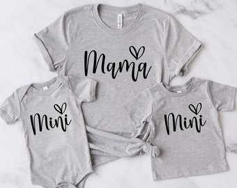 May include: A set of three gray t-shirts and a onesie. The adult shirt says "Mama" with a heart, and the children's shirts and onesie say "Mini" with a heart. All items are made of soft material.