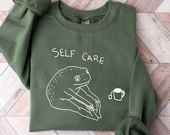May include: Green sweatshirt with a white frog illustration and the text "SELF CARE" printed on the front.