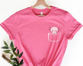 May include: Pink t-shirt with a pocket featuring a cartoon axolotl. The axolotl is pink with black eyes and is peeking out of the pocket.