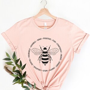 May include: A pink t-shirt with a black bee graphic and the words "Wild, Happy, Smart, Brave, Confident, Fun, Positive, Kind, Humble, Joyful, Creative, Yourself" in a circle around the bee.