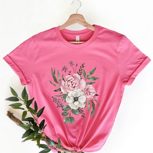 May include: A pink t-shirt with a floral print featuring pink, white, and green flowers. The flowers are arranged in a bouquet shape and are centered on the shirt.