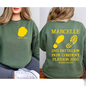 May include: Green sweatshirt with the text "PROUD AUNT" and a yellow footprint on the front. The back of the sweatshirt has the text "MARCELLE", "2ND BATTALION", "PAPA COMPANY", "PLATOON 2093", and "PARRIS ISLAND" along with two yellow footprints.