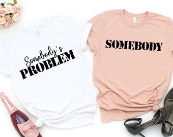 May include: Two t-shirts with a white background and a pink background. The white t-shirt has the text "Somebody's PROBLEM" in black. The pink t-shirt has the text "SOMEBODY" in black.