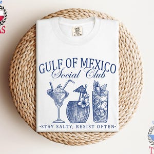 May include: White t-shirt with a blue graphic that reads "Gulf of Mexico Social Club" and features illustrations of cocktails. The phrase "Stay Salty, Resist Often" is printed below the illustrations. The shirt is folded on a woven, circular surface.