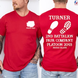 May include: Red t-shirt with white text and graphics. The front of the shirt says "PROUD DAD". The back of the shirt has the name "TURNER", footprints, and text that reads "2ND BATTALION PAPA COMPANY PLATOON 2093 PARRIS ISLAND".
