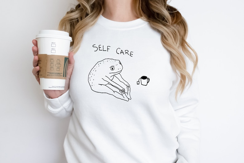 May include: White sweatshirt with a black line drawing of a frog doing yoga and the text "SELF CARE".