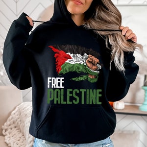 May include: Black hooded sweatshirt featuring the text "FREE PALESTINE" in white and green. The design includes a graphic of the Palestinian flag and a clenched fist. The sweatshirt has a front pocket and a drawstring hood.