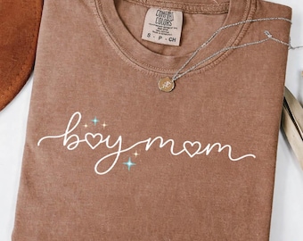 May include: A brown short-sleeve t-shirt featuring the white cursive text "boy mom" with heart accents and small blue star graphics. The shirt has a crew neckline and a visible interior tag that reads "Comfort Colors" and "S P CH".