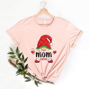 May include: A pink t-shirt featuring a gnome wearing a red hat with white trim and lights. The gnome has a green beard and is holding a candy cane. The text "The Mom Gnome" is printed below the gnome.
