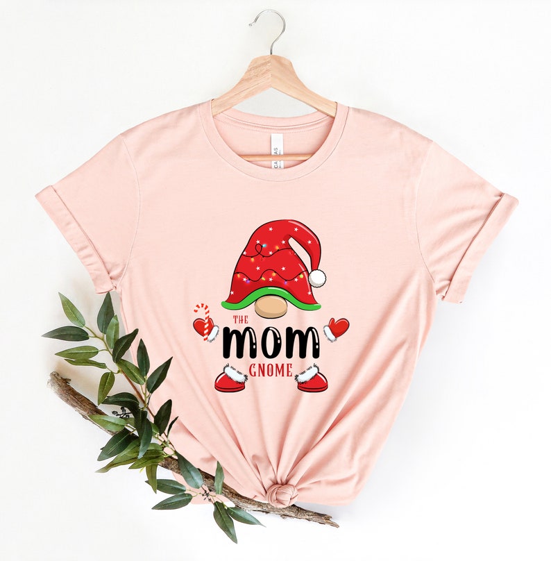 May include: A pink t-shirt featuring a gnome wearing a red hat with white trim and lights. The gnome has a green beard and is holding a candy cane. The text "The Mom Gnome" is printed below the gnome.