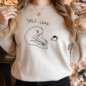 May include: A cream-colored sweatshirt with the text "SELF CARE" printed on the front. The sweatshirt features a black and white illustration of a frog doing yoga with a cup of tea.