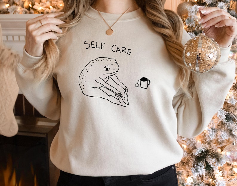 May include: A cream-colored sweatshirt with the text "SELF CARE" printed on the front. The sweatshirt features a black and white illustration of a frog doing yoga with a cup of tea.
