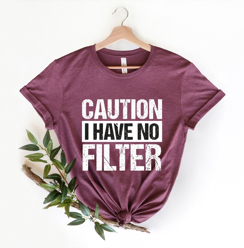 May include: A maroon t-shirt with the text "CAUTION I HAVE NO FILTER" in a distressed white font. The shirt is hanging on a wooden hanger, with a sprig of greenery to the side.