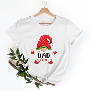 May include: White t-shirt featuring a red and green gnome design. The gnome wears a red hat with white trim and lights. The text "The Dad Gnome" is printed below the gnome.