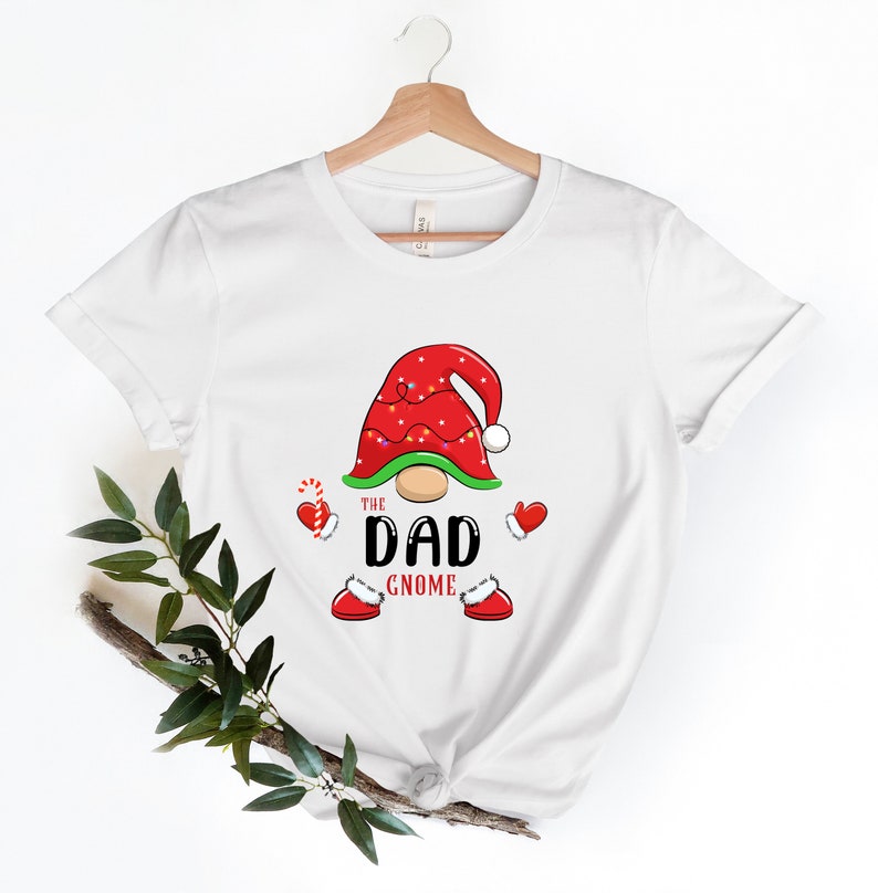 May include: White t-shirt featuring a red and green gnome design. The gnome wears a red hat with white trim and lights. The text "The Dad Gnome" is printed below the gnome.