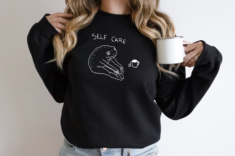 May include: Black sweatshirt with a white graphic of a frog sitting with a cup of tea and the text "SELF CARE".