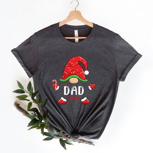 May include: A dark grey t-shirt featuring a Christmas gnome design. The gnome is wearing a red hat with white trim and has a white beard. The text "The Dad Gnome" is printed on the shirt.