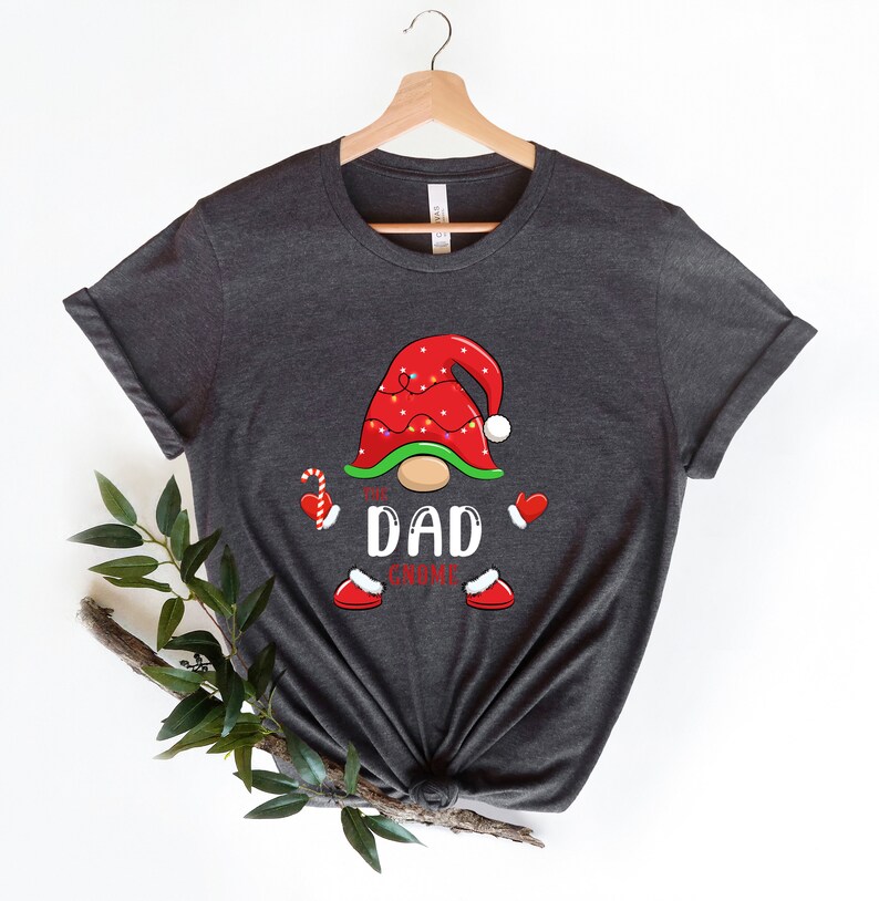 May include: A dark grey t-shirt featuring a Christmas gnome design. The gnome is wearing a red hat with white trim and has a white beard. The text "The Dad Gnome" is printed on the shirt.