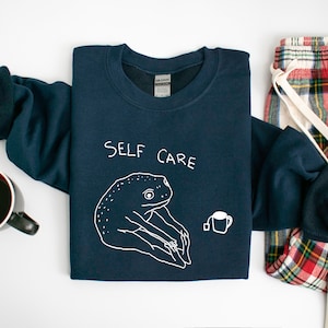 May include: A navy blue sweatshirt with a white graphic of a frog holding a cup of tea. The text "SELF CARE" is printed above the frog.