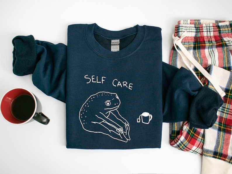 May include: A navy blue sweatshirt with a white graphic of a frog holding a cup of tea. The text "SELF CARE" is printed above the frog.