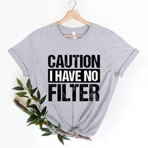 May include: A light gray t-shirt with the text "CAUTION I HAVE NO FILTER" in black and white. The shirt is hanging on a wooden hanger. Green leaves and a branch are placed on the left side of the shirt.