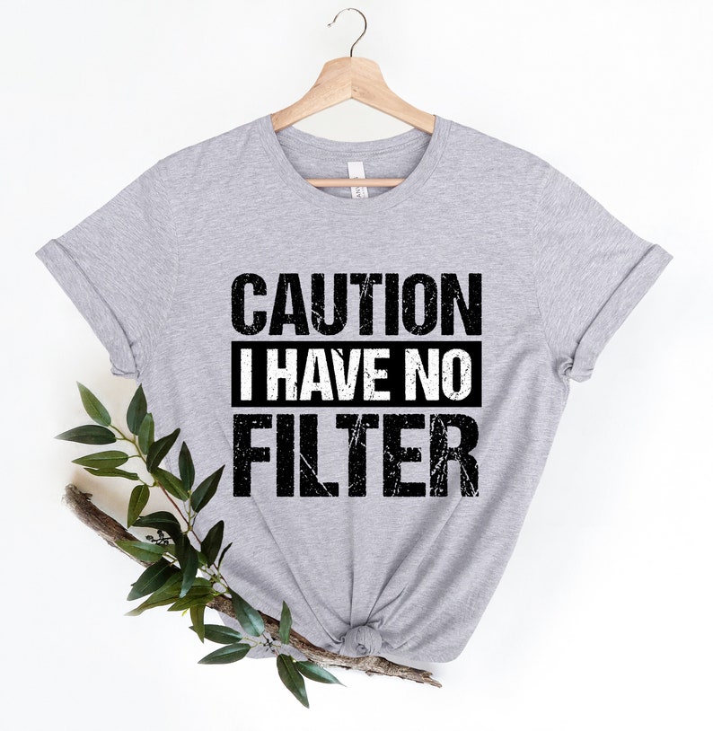 May include: A light gray t-shirt with the text "CAUTION I HAVE NO FILTER" in black and white. The shirt is hanging on a wooden hanger. Green leaves and a branch are placed on the left side of the shirt.