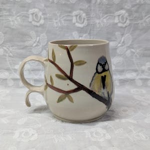 May include: A cream-colored ceramic mug featuring a hand-painted illustration of a blue tit bird perched on a brown tree branch with green leaves. The mug has a unique double-loop handle design and a rounded body shape with a smooth finish.