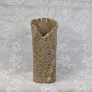 May include: A cylindrical ceramic vase with a speckled beige finish, featuring an uneven, wavy top rim and a textured surface with a vertical row of small perforations and a subtle embossed grid pattern on the side of the vessel.